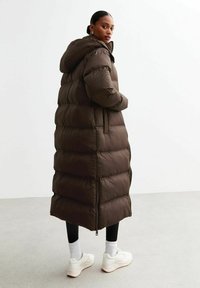 Brun pufferjacka med huva, med stora quiltade sektioner och en helängdsdesign. Bärs över svarta byxor och vita sneakers.