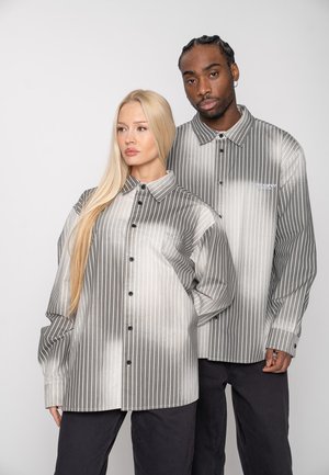 Două persoane care poartă cămăși supradimensionate cu dungi gri și albe, cu nasturi, și pantaloni negri, stând apropiate una de cealaltă.