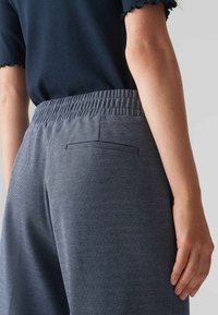 Pantalons tissés gris avec ceinture élastique, dotés d'une poche arrière unique avec un subtil détail de couture. Texture lisse, coupe ajustée.