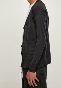 Svart cardigan med knappstängning, ribbade manschetter, två framfickor, lätt material, rund halsringning, bärs över en vit skjorta.