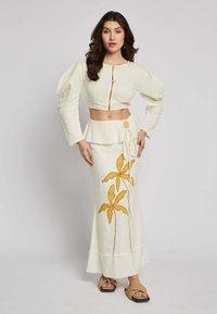 White cropped top with gold zipper, puff sleeves; matching white maxi skirt with yellow leaf embroidery and drawstring waist detail.