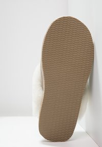 Pantofola color crema con una parte superiore morbida e soffice, dotata di una suola in gomma testurizzata con un motivo ondulato, mostrata dalla vista inferiore.