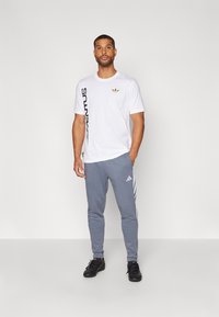 Camiseta blanca de manga corta con texto vertical negro y un logo dorado en el pecho, combinada con pantalones de chándal gris ajustados que tienen rayas blancas.