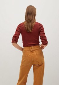 Nainen, jolla on pitkät ruskeat hiukset, pukeutuneena punaisiin ribbivaatteisiin ja korkean vyötärön sinapinvärisiin housuihin, seisoo kädet lanteilla, kasvot pois päin.