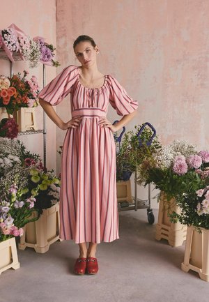 Žena v růžových pruhovaných šatech a červených dřevácích stojí mezi různými květinami v béžových bedýnkách před texturovanou růžovou zdí.