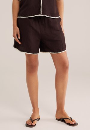 Žena v čiernych šortkách s bielym lemom a zodpovedajúcim topom, stojaca bosá v čiernych žabkách na neutrálne pozadí.