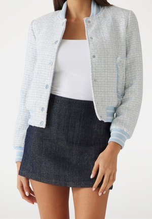 Mujer con chaqueta azul clara texturizada con botones, camiseta blanca sin mangas y minifalda azul oscuro, de pie frente a un fondo blanco liso.