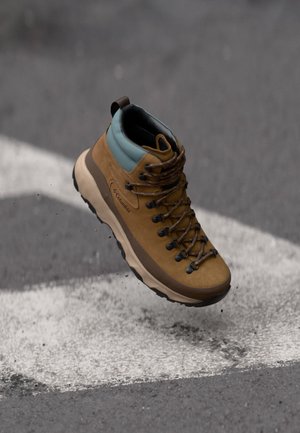 Bota de senderismo de gamuza marrón con un cuello azul claro, cordones marrones y herrajes negros. Presenta una suela intermedia blanca gruesa y una suela exterior texturizada negra.