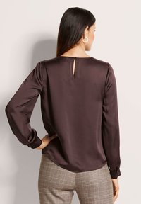 Frau mit langen dunklen Haaren, die eine dunkelbraune Satinbluse mit Knöpfen im Rücken und Karohosen trägt, steht mit einer Hand auf der Hüfte.