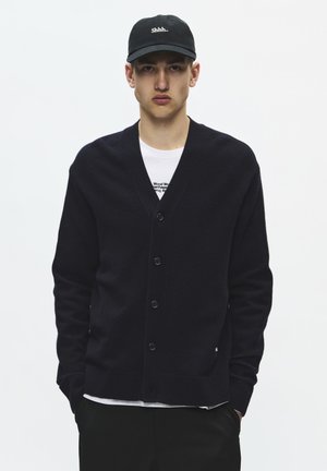 Jeune homme portant un cardigan noir boutonné sur une chemise blanche et une casquette noire avec le texte "Shhh..", debout les mains dans les poches.