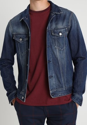 Hombre con chaqueta vaquera azul oscuro sobre una camiseta burdeos y pantalones azul marino con estampado de cuadros rojos, con las manos en los bolsillos.