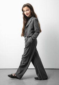 Szary garnitur w paski z krótką marynarką i szerokimi spodniami. Teksturowana tkanina, w zestawieniu z czarnymi butami z ćwiekami. Długie włosy stylizowane luźno.