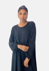 Vestido negro de manga larga con cuello redondo, cintura fruncida y textura suave. Modelo acompañado de pendientes y pulseras de oro.