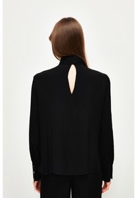 Blusa nera a maniche lunghe con colletto alla cinese e apertura a goccia sulla schiena. Presenta polsini con bottoni e una texture liscia.