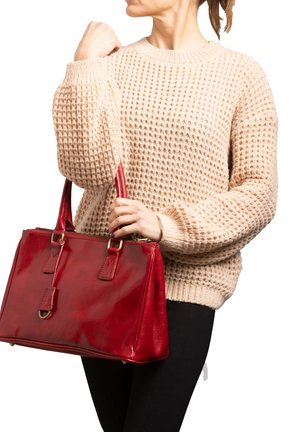 Borsa a tracolla in pelle rossa con finitura lucida, accessori in oro e una forma strutturata. Un maglione intrecciato beige contrasta con il colore audace della borsa.