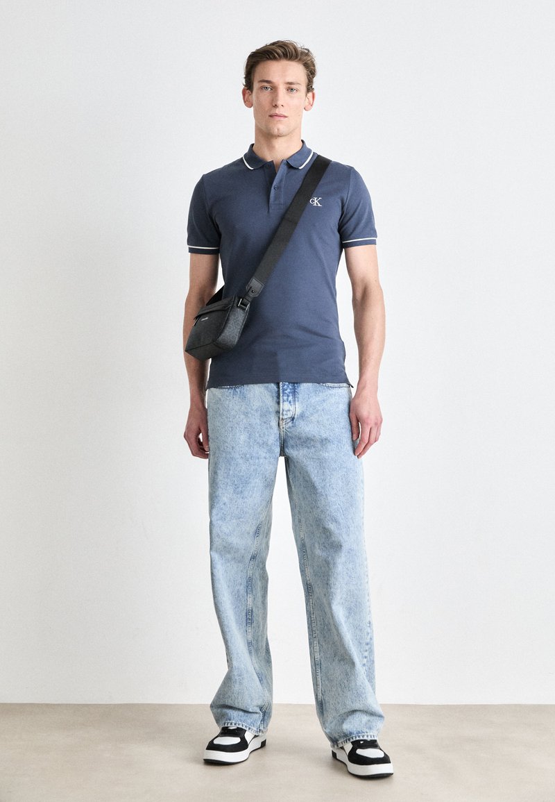 Marineblaues Polohemd mit weißen Akzenten, hellblaue weite Jeans, schwarz-weiße Sneakers und eine graue Umhängetasche.