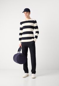 Dryžuotas megztinis su nėriniais kreminiu ir tamsiai mėlynu atspalviu, juodos kelnės, baltos sportinės bateliai, laiko apvalią tamsiai mėlyną sportinę rankinę ir tamsiai mėlyną kepurę su logotipu.