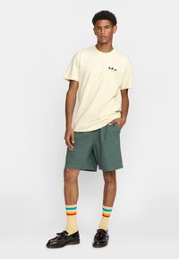 Beige katoenen t-shirt met een teal berglogo, groene shorts en multicolore gestreepte sokken. Zwarte schoenen met een glanzende afwerking.