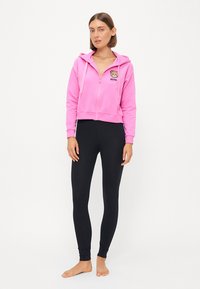 Roza kratka kapuca s prednjim zadrgo in grafiko medveda, v kombinaciji s črnimi oprijetimi leggingi. Gladka tekstura, sproščen oblikovanje in minimalno blagovno znamčenje.