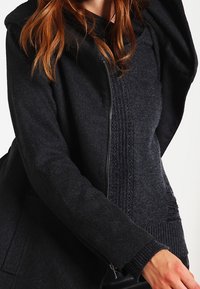 Chaqueta negra con cremallera y panel de punto texturizado, que incluye capucha y puños ajustados para mayor calidez y comodidad.