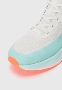 Valge sportlik jalanõud tekstureeritud pealispinnaga, piparmündiroheline aktsent ja oranž kand. Omab kerget materjali ja detailset tikandit.