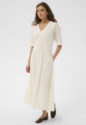 Kobieta o długich kręconych włosach, ubrana w kremową sukienkę midi z ażurowym wzorem i dekoltem w serek, z rękawami do łokci oraz czarne buty na niskim obcasie, stojąca z jedną ręką w kieszeni.