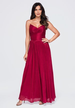 Vrouw met lang donker haar die een strapless, tot op de vloer reikende rode jurk draagt en zilveren sandalen met hoge hakken, poseert met één hand op haar heup.