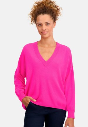 Pull rose en V à manches longues et coupe décontractée, fabriqué en tissu doux, avec bords-côtes aux poignets et à l'ourlet. Porté par-dessus un pantalon bleu marine.