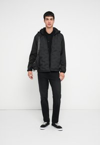 Svart dunjakke med hette, kombinert med en svart polotrøye og mørke jeans. Modellen har på seg svarte sneakers og bærer en liten veske.