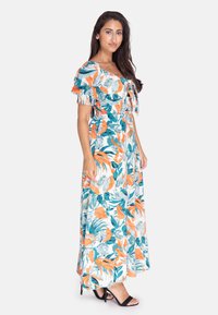 Blomstret maxikjole i hvid med orange og teal bladmønstre. Har flæser påærmerne, en tætsiddende bodice og en flydende silhuet.