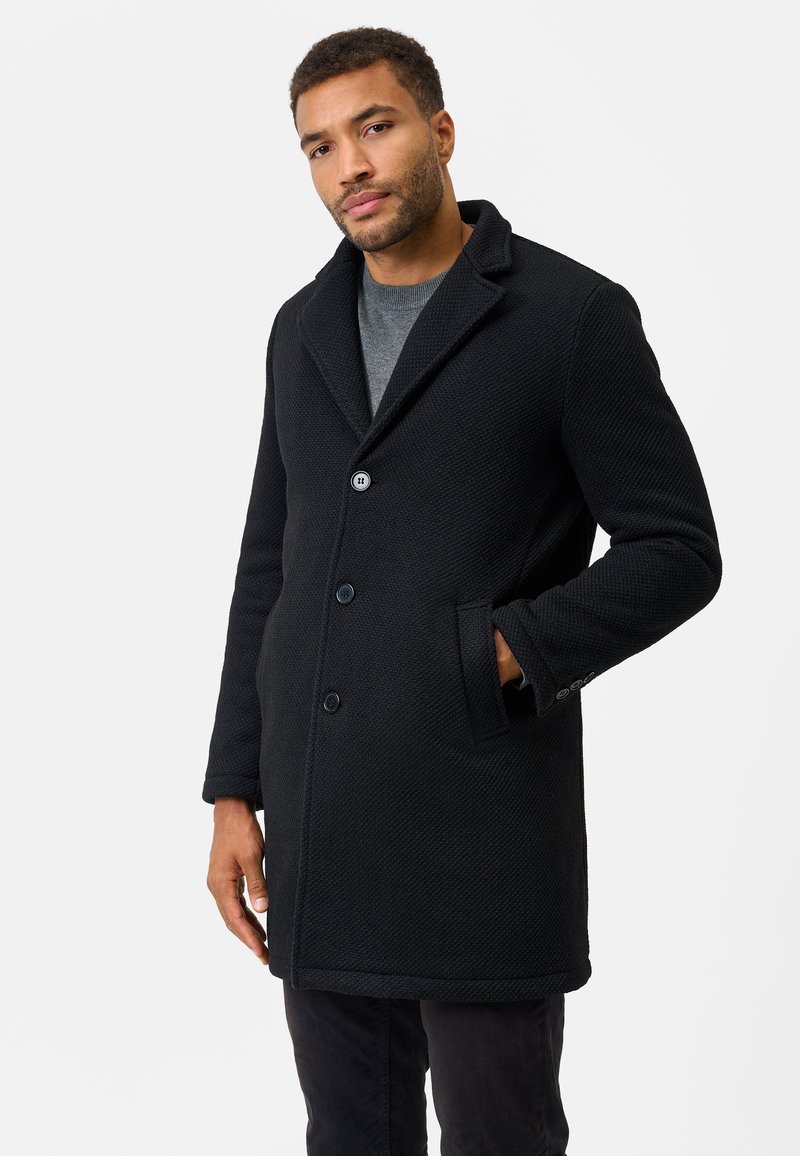 Sort tekstureret frakke med en stor krave, tre knapper og sidelommer. Båret over en grå sweater, som har en lige silhuet.