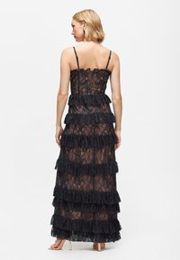 Černá maxi šaty z krajky s vrstvenými volány, tenkými nastavovacími ramínky a průhledným designem, který odhaluje jemný kontrast textur.