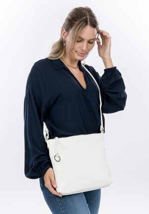 Frau mit hellbraunen Haaren, die eine dunkelblaue Bluse und Jeans trägt und eine weiße Umhängetasche mit einem silbernen Reißverschlussring hält.