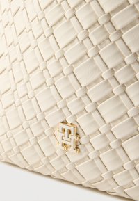 Kremowa torba z plecionej skóry o gładkiej teksturze, wzorze w kratę oraz złotym akcentem logo w dolnym rogu.