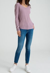 Fialový pletený svetr s výstřihem do V a dlouhými rukávy, kombinovaný s modrými džínami a bílými teniskami. Hladká textura a uvolněný střih.