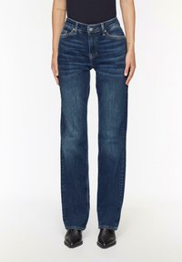 Jeans en denim bleu avec une coupe droite, lavage moyen, cinq poches et éléments en métal. Portés avec des bottines noires.