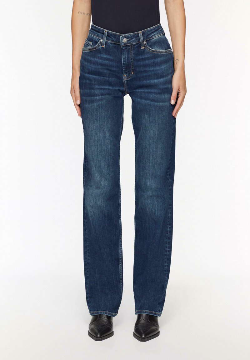Jeans en denim bleu avec une coupe droite, lavage moyen, cinq poches et éléments en métal. Portés avec des bottines noires.