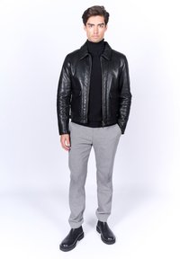 Chaqueta de cuero negra con cremallera, dos bolsillos delanteros y un cuello, llevada sobre un cuello de tortuga negro. Pantalones grises con estampado y botas negras.