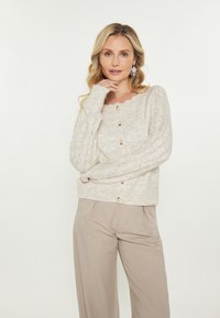 Cardigan lavorato a maglia color beige chiaro con scollatura a onde, bottoni in legno e dettagli a coste sulle maniche, abbinato a pantaloni beige.