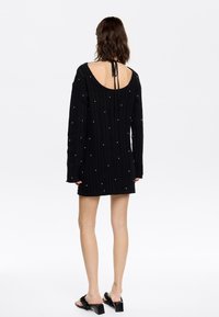 Schwarzes gestricktes Kleid mit langen Ärmeln, offenem Rücken und Rückenknoten-Detail. Verfügt über kleine dekorative Akzente überall. Kurze Länge.