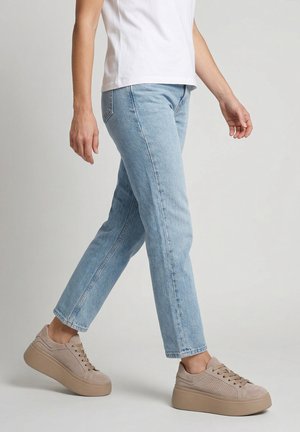 Osoba ubrana w jasnoniebieskie dżinsy, beżowe sneakersy na platformie i białą koszulkę, robi krok na jednolitym jasnym tle.