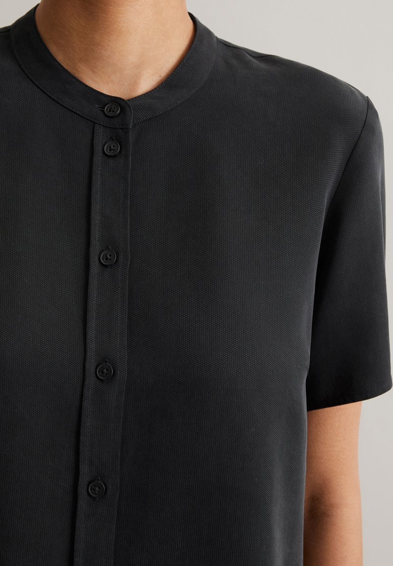 Schwarzes Kurzarmhemd mit Kragen, Knopfverschluss und strukturiertem Stoff. Das Design besticht durch einen klaren und minimalistischen Stil.