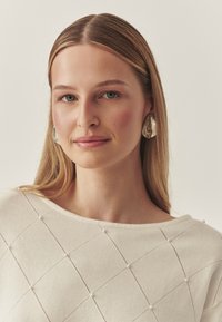 Tejfehér pulóver gyémántmintával és apró díszítésekkel, széles nyakkivágással. Ezüst színű, figyelemfelkeltő fülbevalók teszik teljessé a megjelenést.