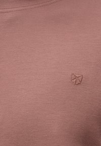 Κοντινή λήψη ενός ροζ υφάσματος με μια μικρή, ραμμένη γεωμετρική λογότυπη κοντά στο πάνω κέντρο του.