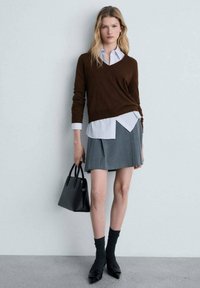 Brauner Pullover über einem weißen Hemd mit Kragen, grauer plissierter Rock, schwarze Handtasche, kniehohe Socken und spitze schwarze Schuhe.
