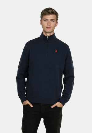 Marineblauer Pullover mit Viertelreißverschluss und langen Ärmeln, ausgestattet mit einem kleinen roten Logo auf der Brust und gerippten Bündchen. Glatte Stoffoberfläche.