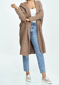 Tan knitted cardigan with a hood, open front, and ribbed cuffs, paired with light blue straight-leg jeans and beige sneakers.