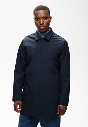 Homme portant un manteau mi-long bleu marine foncé par-dessus un haut bleu zippé sur un fond blanc uni.