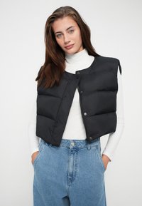 Vrouw in een witte coltrui, zwarte gewatteerde vest en hoogtijde blauwe jeans met handen in haar zakken voor een effene achtergrond.