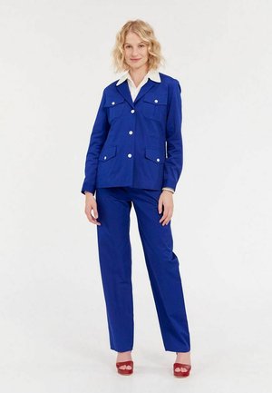 Veste bleue avec deux poches avant, boutons blancs et col blanc, associée à un pantalon bleu assorti et des sandales à talons rouges.
